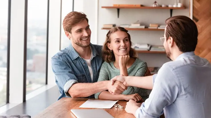 Ein Pärchen sitzt einem Mann gegenüber und die Männer schütteln sich die Hand. Alle haben entspannte Gesichtszüge was die vertrauensvolle Beziehung zwischen Mietende und Vermieter darstellen soll.